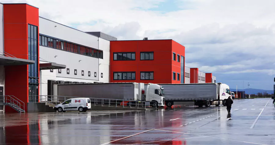 Photo of a red and grey industrial warehouse with a rainy reflective parking lot in front