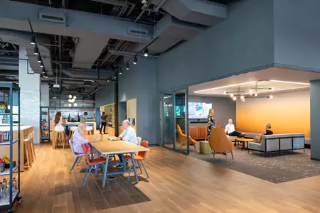 Open plan layout office hallway with tables and corner gathering space with couches and a television